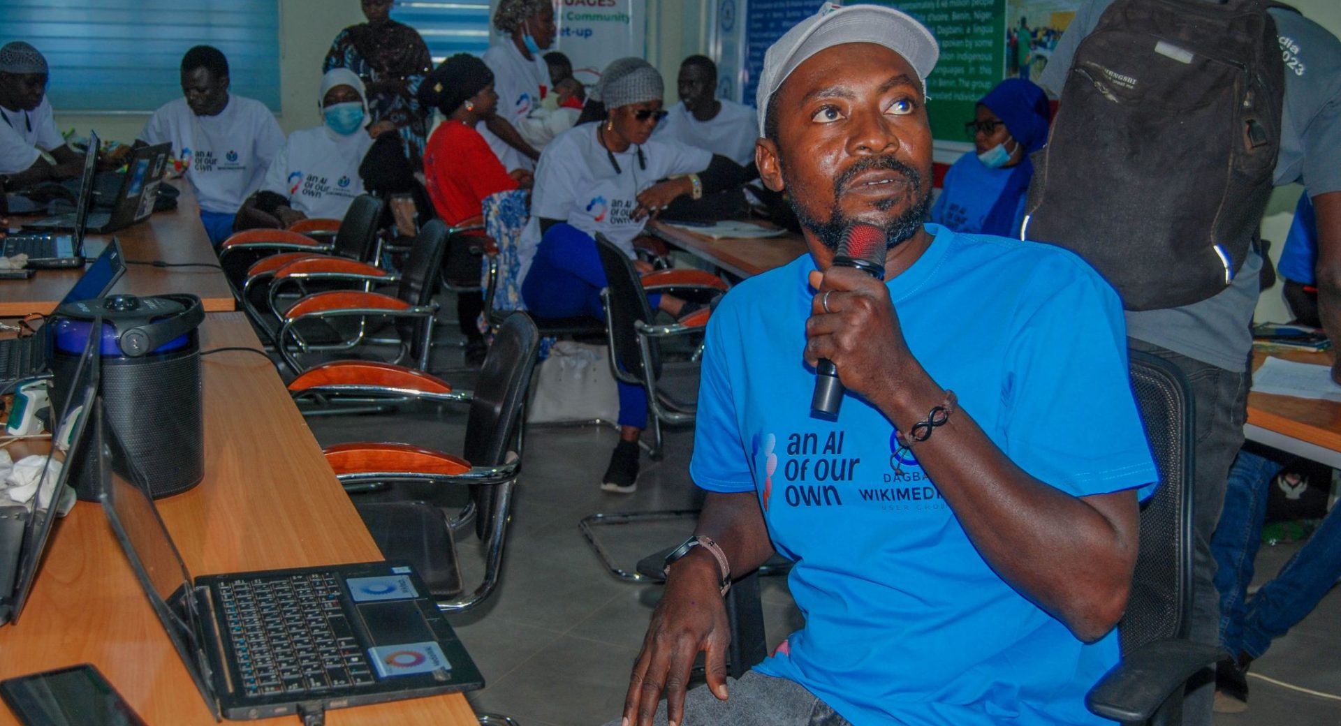 A man in an AAOO shirt holding a mic and looking to the right in a classroom with others looking on